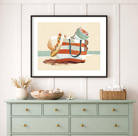 Beach bag filled with towel, sunglasses, and sun hat on the sand with ocean in the background, coastal lifestyle art print displayed in a beach house interior