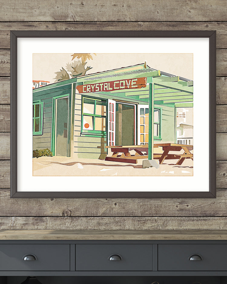 Framed artwork of a beachside building with 'Crystal Cove' sign on a wooden wall.