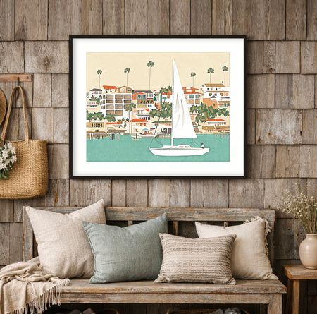 Sailboat moving through a coastal channel with bluffs, beach houses, and palm trees in the background, coastal art print displayed in a beach house interior