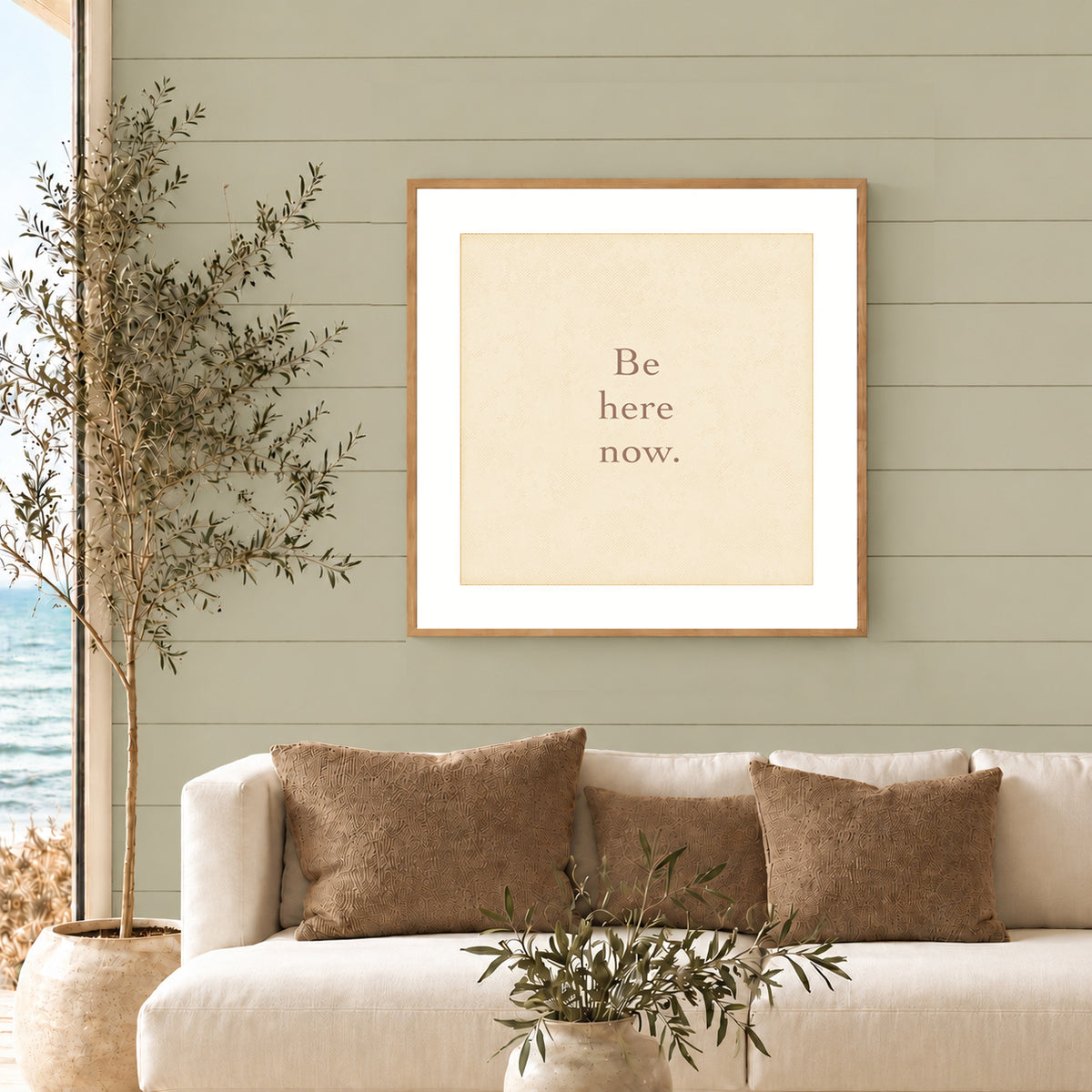 Coastal living room with a square framed ‘Be here now.’ art print above a white sofa, against a muted sage green paneled wall.