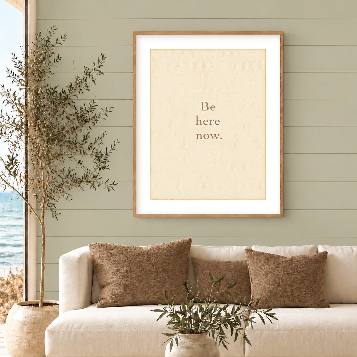 Coastal living room with a vertical framed ‘Be here now.’ art print above a white sofa, against a muted sage green paneled wall.