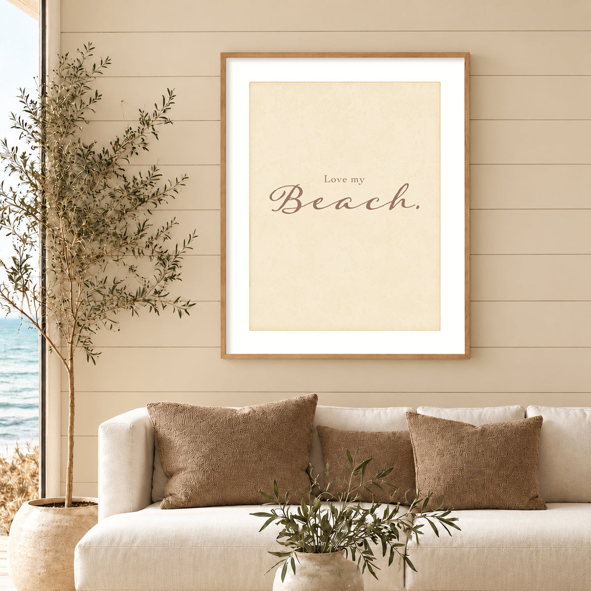 Coastal living room with a framed ‘Love my Beach.’ vertical art print above a cream sofa, featuring a muted sage green shiplap wall, tan pillows, and a potted olive tree beside an ocean view.