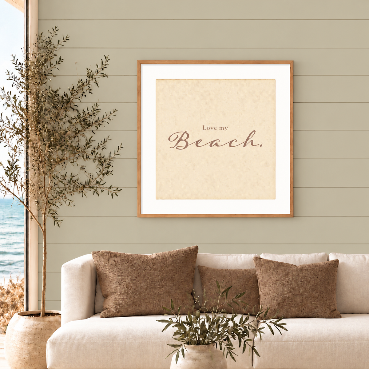 Coastal living room with a framed ‘Love my Beach.’ square art print above a cream sofa, featuring a muted sage green shiplap wall, tan pillows, and a potted olive tree beside an ocean view.